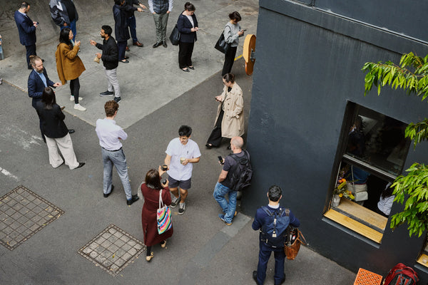 An overhead view of a group of people standing and walking on a paved outdoor area beside a Patricia 1 with a window and green leaves at the edge.