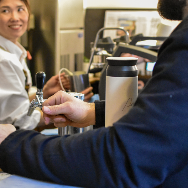 A cream coloured Kinto Tumbler with the Patricia logo, sitting on the counter in front of a bearded gentleman. A smiling barista steams milk in the background.