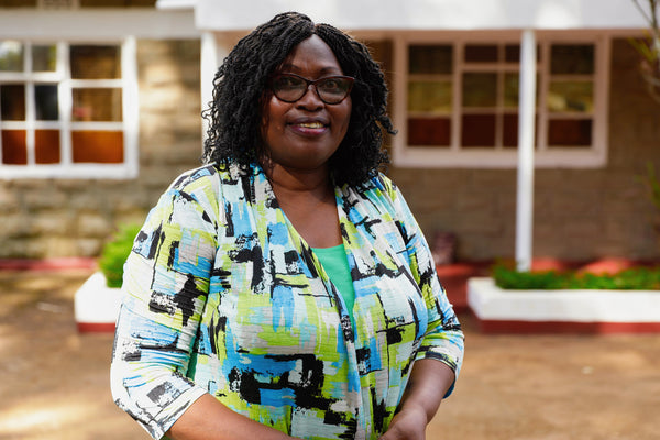 Lucy Wanja Ndogo smiling and wearing a colourful shirt, in front of the Rurima homestead.