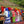 A group of people in colorful traditional clothing and headscarves stand along wooden tables outdoors, sorting red coffee cherries in a rural setting with a grassy hill and trees in the background.