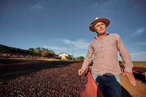 Jean Vilhena wearing a wide-brimmed hat, a red and white checkered long-sleeve shirt, and blue jeans stands outdoors in a farm setting next to a raised bed of dark red coffee cherries under a clear blue sky.