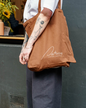 A person wearing a white shirt and dark blue pants carries a brown canvas tote bag with white 'Patricia Coffee Brewers' branding against a dark wall.
