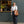 A person stands outdoors in front of Patricia Lt Bourke St, wearing a white short-sleeve button-up shirt, dark wide-leg trousers, and carrying a large brown tote bag with the Patricia logo.