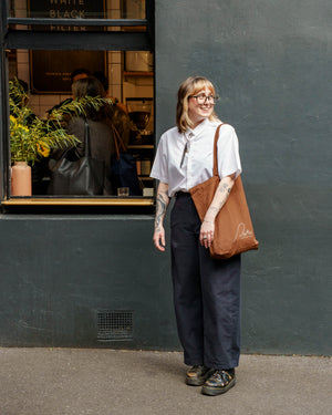 A person stands outdoors in front of Patricia Lt Bourke St, wearing a white short-sleeve button-up shirt, dark wide-leg trousers, and carrying a large brown tote bag with the Patricia logo.