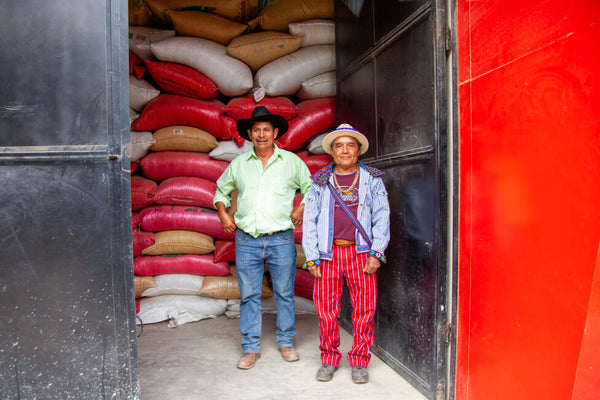 El Sendero Cooperative manager Pablo Gaspar with coffee producer and member Feliciano. They stand in front of stacks of pink, orange and white coffee bags.