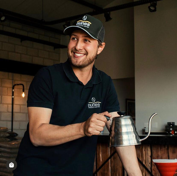 Gabriel Nunes wearing a Nunes branded cap and shirt, smiling and holding a coffee kettle in a kitchen setting.