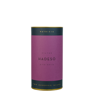 A navy blue cylinder with a purple label describes the roasted coffee beans within. 'PATRICIA' and 'TWO HUNDRED GRAMS' are printed in gold foil.