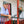 Jairo Arcila stands on a red balcony wearing a shirt and green cap, smiling broadly and holding a small yellow fruit.