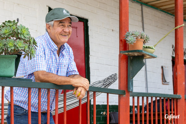 Jairo Arcila stands on a red balcony wearing a shirt and green cap, smiling broadly and holding a small yellow fruit.