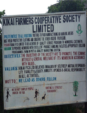 A large white sign with black and blue text reading 'KIKAI FARMERS COOPERATIVE SOCIETY LIMITED' and various policies and values, photographed outdoors with trees and sky visible around it.