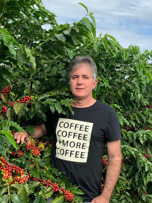 Jean Vilhena standing in a coffee plantation wearing a black t-shirt with a white graphic that reads 'COFFEE COFFEE + MORE COFFEE'.