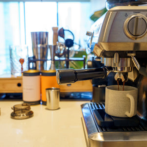 A stainless steel espresso machine extracting coffee into a light gray ceramic mug on a kitchen counter, with coffee beans and other kitchen items in the background.