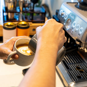 A close-up lifestyle shot of a person pouring milk from a stainless steel pitcher into a gray ceramic mug, positioned next to a modern silver espresso machine.