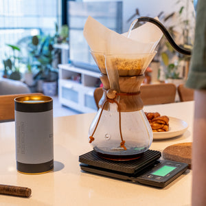 A Chemex pour-over coffee maker with a paper filter and glass carafe sits on a digital scale on a light-colored table, with hot water being poured from a black gooseneck kettle. A person wearing a green shirt is partially visible on the right, and a blue coffee cylinder is on the left.