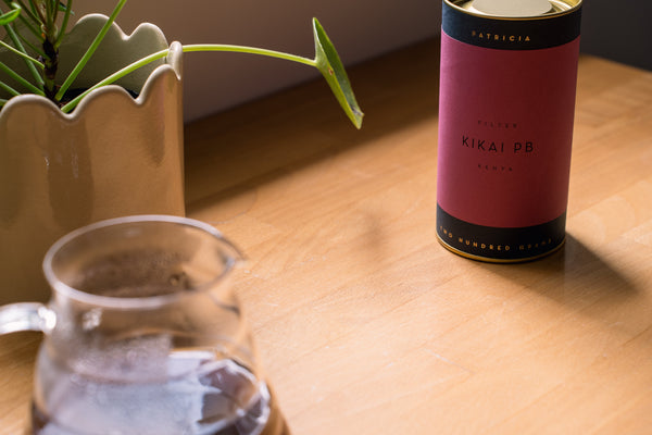 A purple cylindrical coffee tin labeled 'PATRICIA KIKAI PB' sits on a wooden table next to a glass teapot and a potted plant.