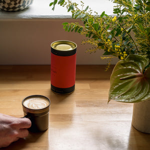 A red coffee cylinder stands on a wooden table beside a cup of white coffee, with a hand holding one mug and a decorative plant in the foreground.