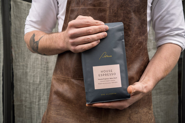 A person wearing a brown leather apron holds a dark blue bag of Patricia House Espresso coffee out front of Patricia Lonsdale St.