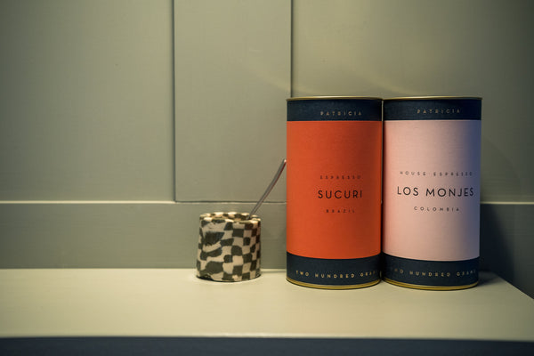 Two cylindrical Patricia coffee tins, one red with 'SUCURI' and one pale pink with 'LOS MONJES', standing on a shelf next to a small chequered cup with a spoon.