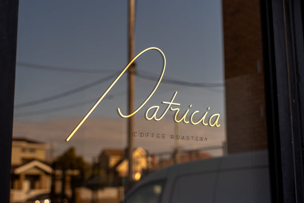 A gold decal sign spelling 'Patricia' with 'COFFEE ROASTERY' underneath, mounted on a window. The glass reflects the outdoor street scene at dawn.