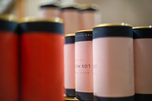 A close-up row of cylindrical tins with pale blush pink labels and dark navy bands, some with gold tops. The foreground tins are in sharp focus while those behind them blur softly due to shallow depth of field.
