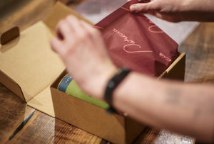 A close-up of a person's hands opening a brown cardboard box on a wooden table, revealing a dark red tissue paper covering green cylindrical coffee tin.
