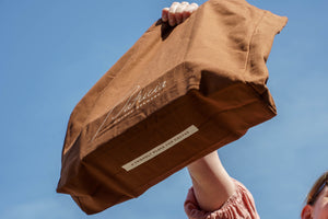 A person outdoors holds up a brown cotton tote bag with white script reading 'Patricia', with 'A FRIENDLY PLACE FOR COFFEE' written on the underside of the bag.