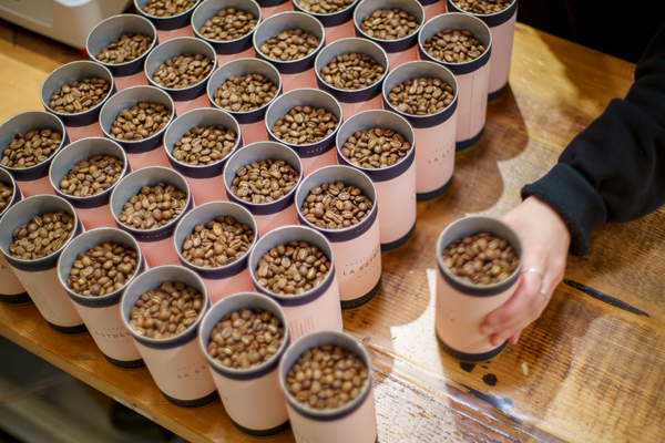 Person arranging 200g cylinders of House Espresso beans beans on a wooden surface