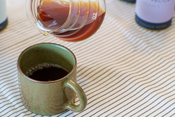 Coffee being poured from a glass pot into a green ceramic mug on a striped fabric surface.