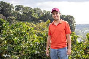 Sebastian Gomez standing in front of coffee trees, wearing an orange shirt and red cap reading 'Cofinet'.