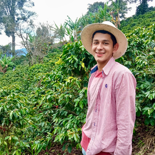 Camilo Londoño smiling, wearing a straw hat and red striped shirt standing among coffee plants